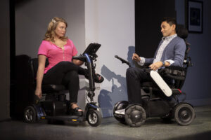 Madison Ferris and Danny Gomez, actors in All of Me, sit across from each other on stage in their power mobility devices, looking at each other and using AAC to communicate.