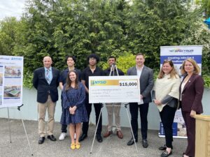 A diverse group of 8 stands in front of trees, smiling, with a large $15,000 check from NYSID to the NYIT design group. To their left is a sign about the silk screen cleaner design and to the right is a Spectrum Designs sign.